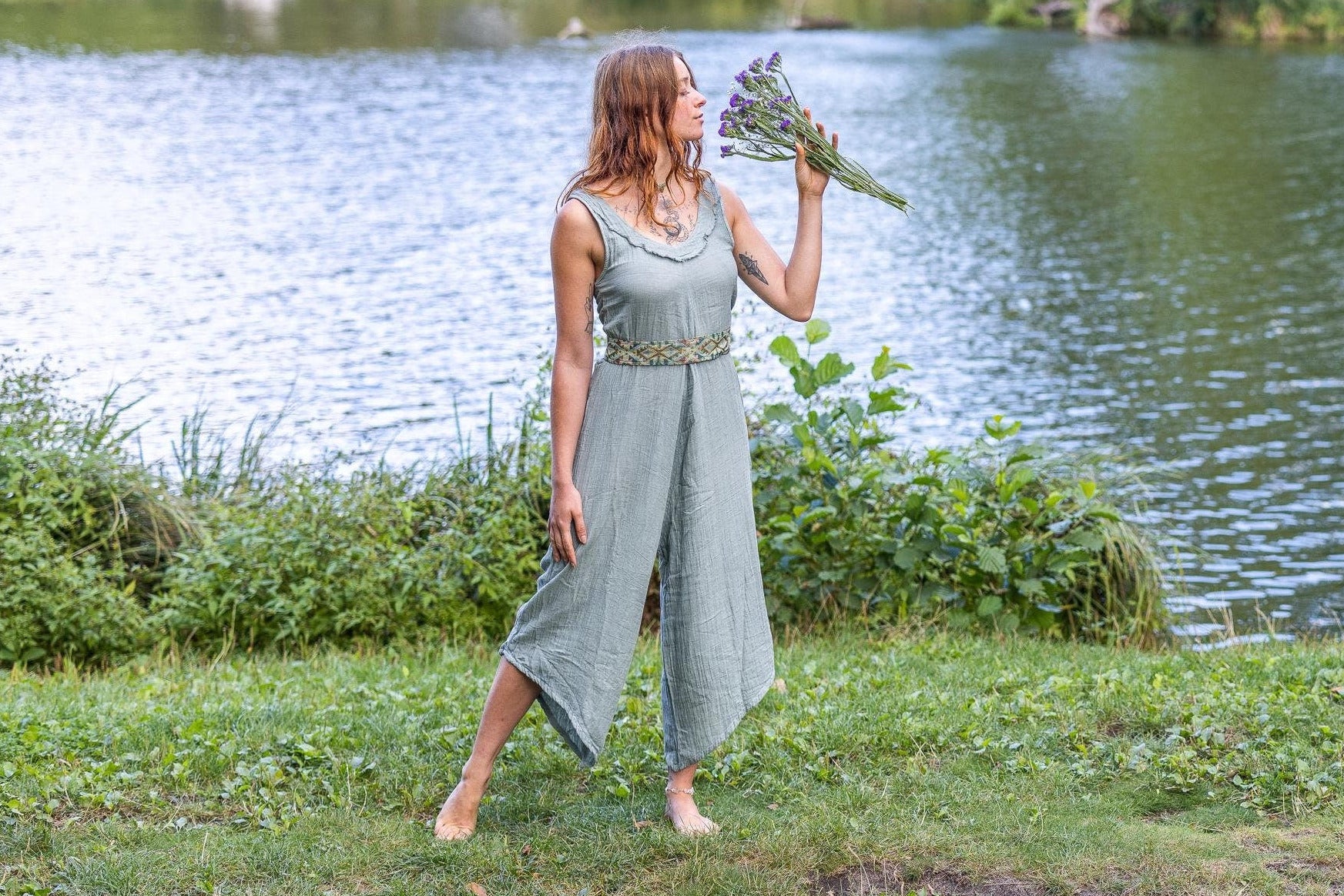 Woman in a sage green jumpsuite with an ethnic belt, standing by a lake holding flowers, surrounded by nature.